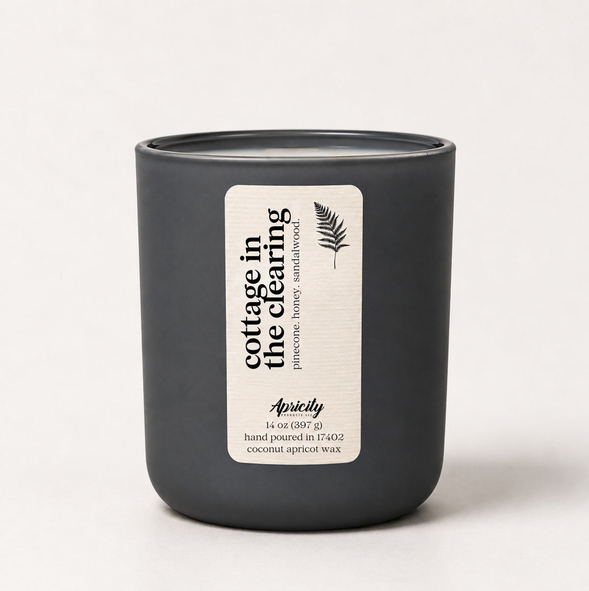 Cottage in the Clearing 14 oz candle in a matte charcoal tumbler with a cream fern label reading pinecone, honey, sandalwood
